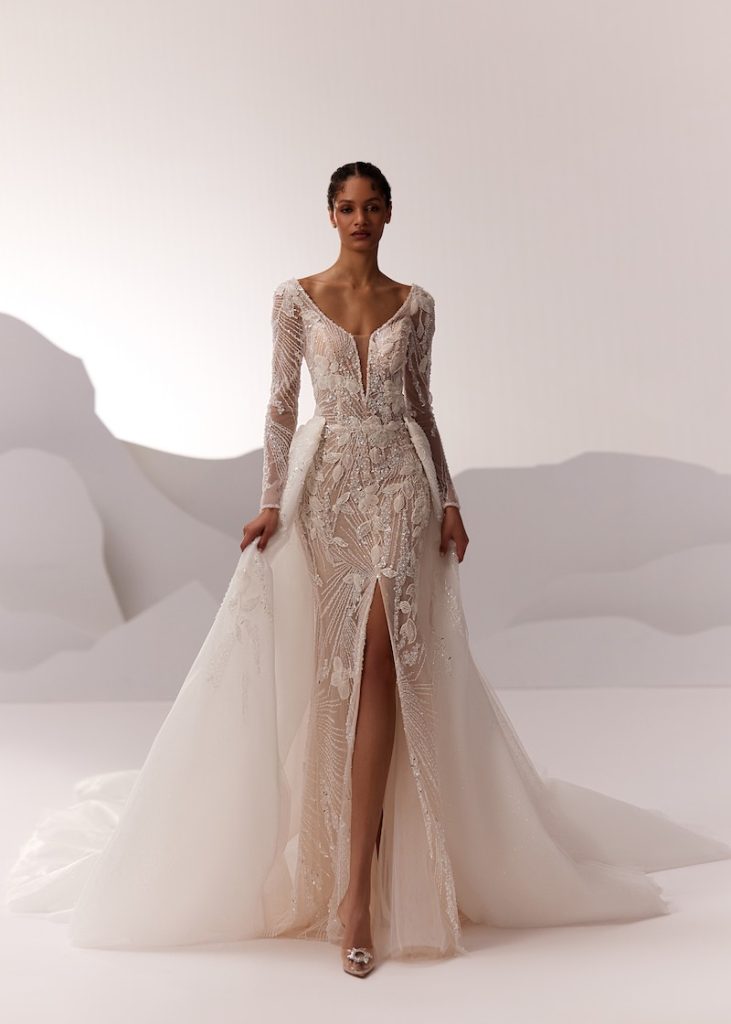 In questa foto, una modella posa  indossando un abito da sposa Elena Morar 2026 modello a sirena con ricami di perline e paillettes con spacco frontale, con maniche lunghe, scollo profondo a V e sovragonna in tulle 