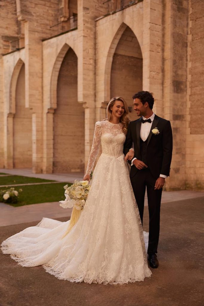 In questa foto, una modella posa con un modello in smoking indossando un abito da sposa Elena Morar 2026 interamente in pizzo, con gonna ad A, bustier e maniche lunghe