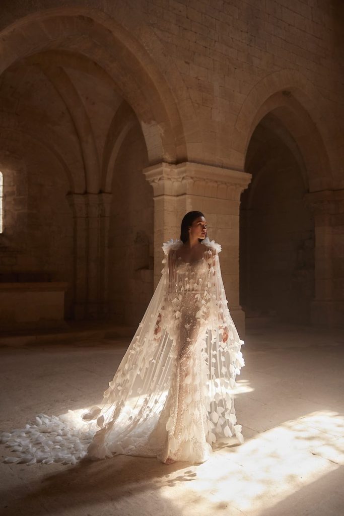 In questa foto, una modella posa  indossando un abito da sposa Innocentia 2026 a sirena con cappa in tulle impreziosita da fiori 3D all over