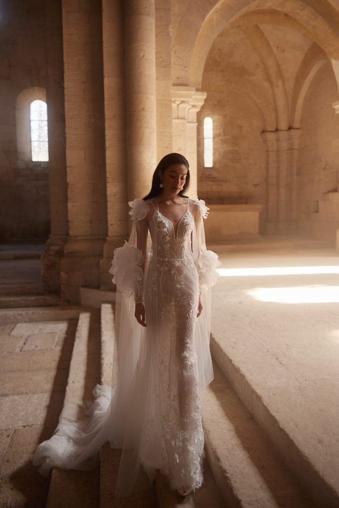 In questa foto, una modella posa  indossando un abito da sposa Innocentia 2026 modello a sirena con ricami di perline e paillettes, mantella in tulle e maxi fiori 3D sulle spalle e all'altezza dei gomiti