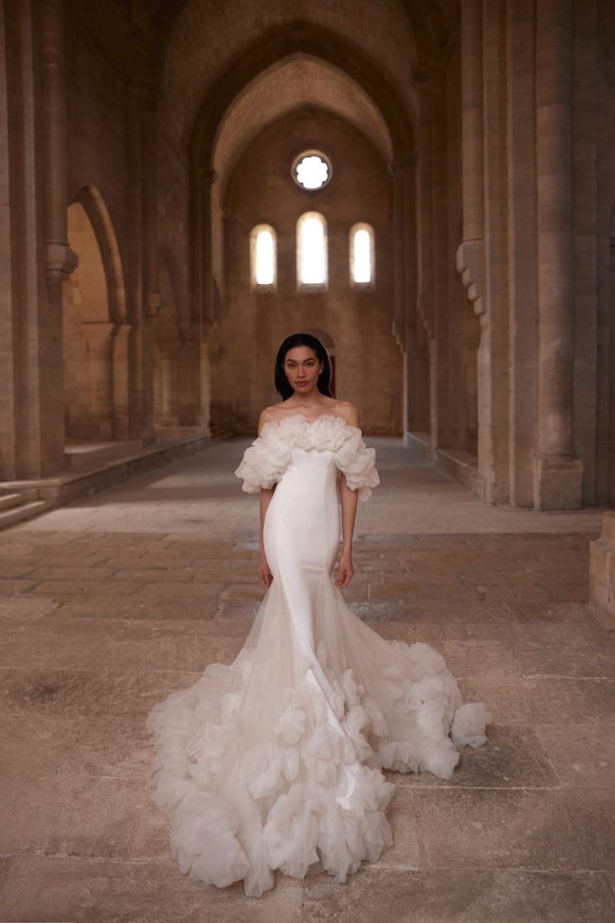 In questa foto, una modella posa  indossando un abito da sposa Innocentia 2026 a sirena in tessuto stretch e rouches sulla scollatura off shoulder e sull'orlo