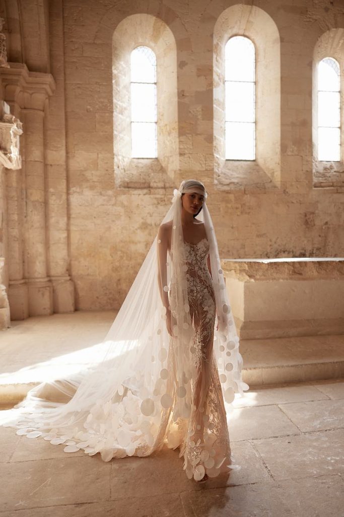 In questa foto, una modella posa  indossando un abito da sposa Innocentia 2026 a sirena effetto nude con ricami 3D. La modella indossa anche un velo stretto sulla testa con motivi circolari sull'orlo