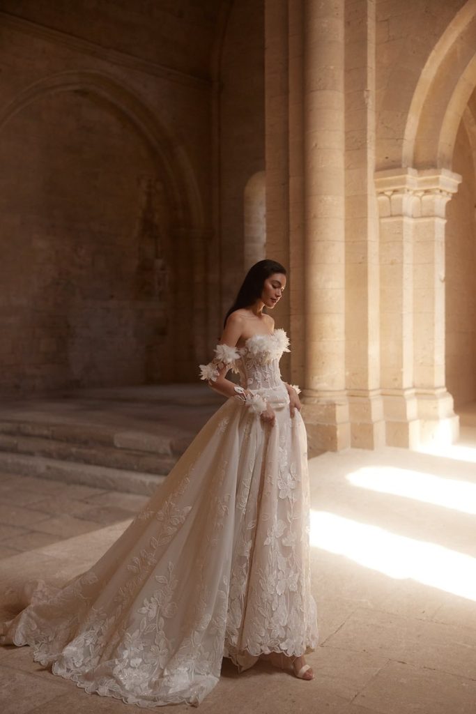 In questa foto, una modella posa  indossando un abito da sposa Innocentia 2026 modello ad A con ricami floreali, bustier e fiori 3D sull'orlo e sui manicotti