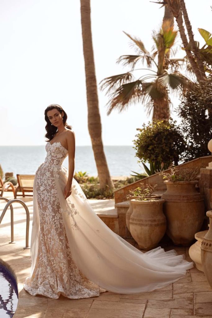 In questa foto, una modella posa  indossando un abito da sposa Perfioni 2026 interamente in pizzo floreale effetto nude con sovragonna in organza decorata 