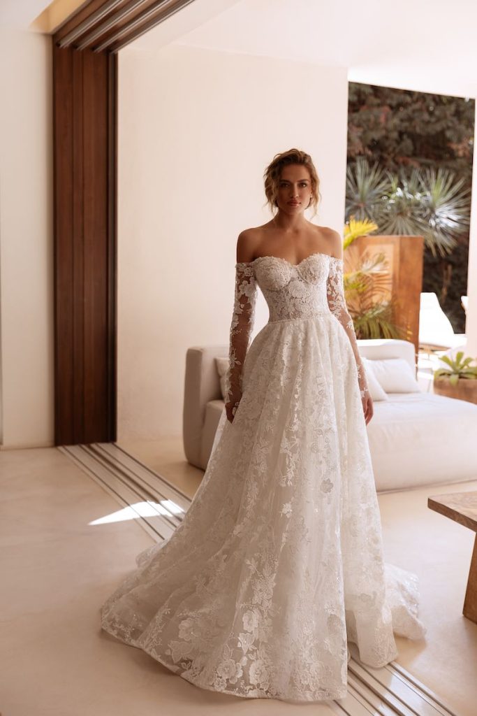 In questa foto, una modella posa  indossando un abito da sposa Perfioni 2026 modello ad A, in pizzo floreale, con scollo a cuore e maniche lunghe 
