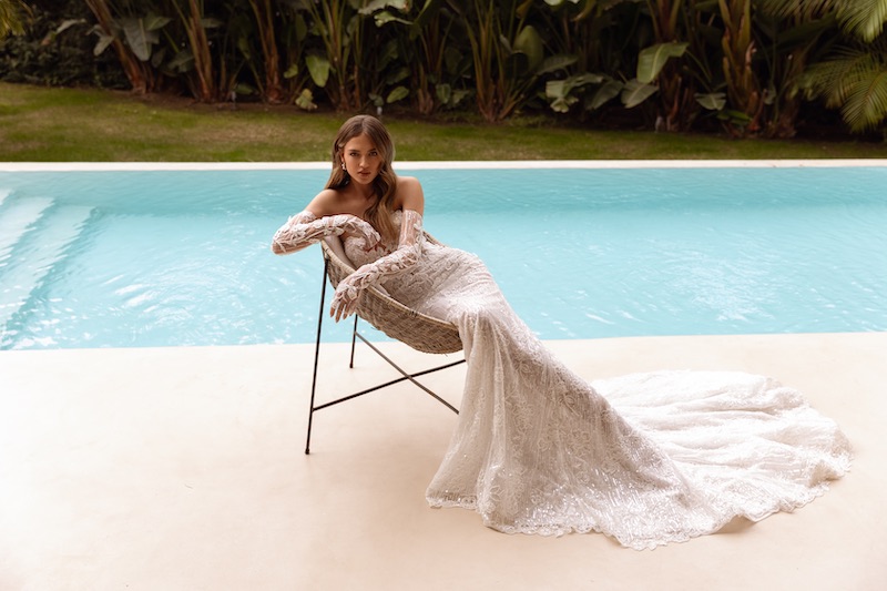 In questa foto, una modella posa seduta su una sedia a bordo piscina indossando un abito da sposa Perfioni 2026 a sirena con lungo strascico, in pizzo con ricami preziosi e manicotti abbinati