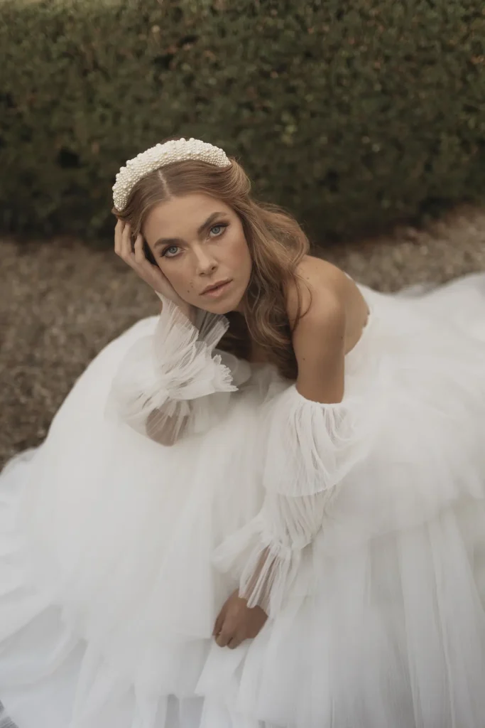 In questa foto di Vincenzo Pioggia si vede una sposa seduta a terra su una superficie di ghiaia, che guarda intensamente verso l’obiettivo. Indossa un abito da sposa vaporoso di colore bianco, con maniche lunghe in tulle trasparente e plissettato. I capelli biondi sono sciolti e ondulati, impreziositi da un cerchietto rigido di colore bianco decorato con perle. Sullo sfondo si intravede una siepe verde, sfocata. L’espressione della sposa è intensa e sognante.