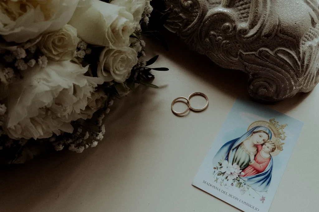 In questa foto di Vincenzo Pioggia si vede una composizione intima e simbolica: due fedi nuziali dorate poggiano su una superficie chiara accanto a un'immaginetta religiosa raffigurante la Madonna del Buon Consiglio con il Bambino Gesù. A sinistra, un bouquet di rose bianche e fiori di gipsofila aggiunge un tocco floreale, mentre in alto a destra si intravede una scultura decorativa in pietra. L’immagine trasmette un senso di sacralità e delicatezza, tipico del momento del matrimonio.