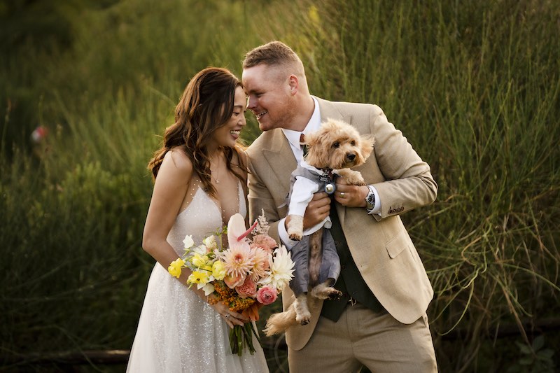 In questa foto di Valeria Santoni, due sposi felici e sorridenti in un prato. Lo sposo tiene in braccio un cagnolino Yorkshire con un vestitino di colore grigio. La sposa tiene in mano un bouquet di fiori colorati