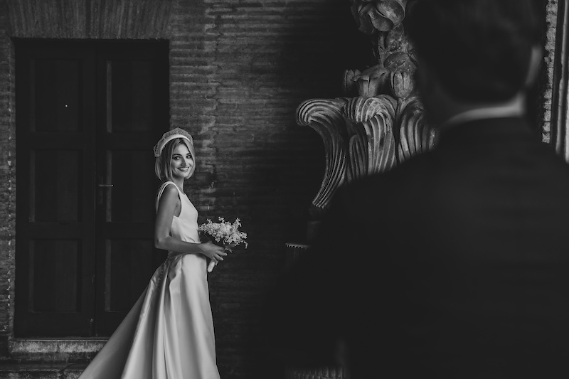 In questa foto in bianco e nero, una sposa sorridente all'arrivo dello sposo ripreso di spalle e fuori fuoco