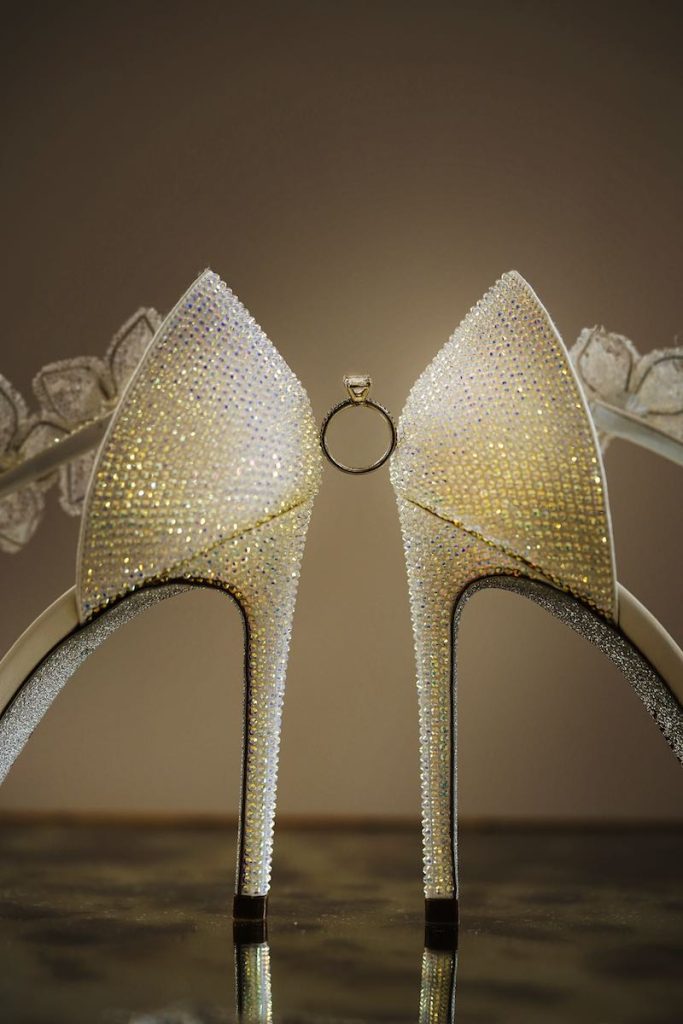 In questa foto, il dettaglio dell'anello di fidanzamento tenuto tra i tacchi di due scarpe da sposa