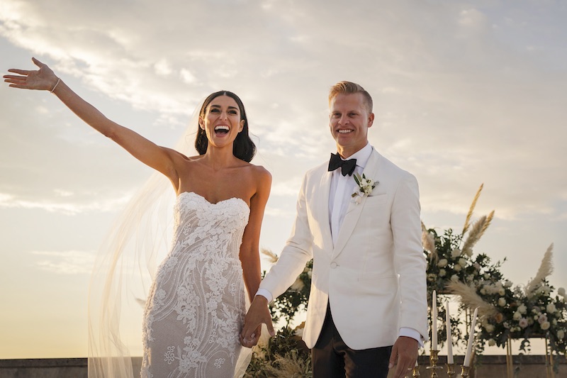 In questa foto di Valeria Santoni, due sposi felici al termine della cerimonia di nozze celebrata al tramonto. la sposa indossa un abito a sirena con ricami floreali e scollo a cuore; lo sposo indossa uno smoking con giacca bianca