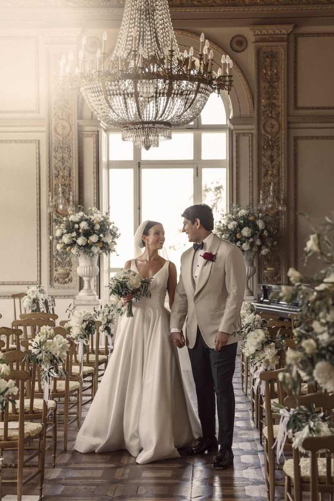In questa foto di Valeria Santoni due sposi si guardano felici mentre camminano verso l'uscita dalla cerimonia in una sala elegante, circondati da fiori bianchi