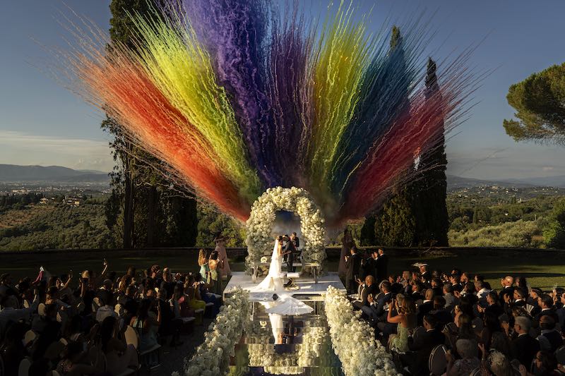 In questa foto di Valeria Santoni, la scena di un bacio tra due sposi al termine della cerimonia di nozze, ripresa dall'alto tra fuochi d'artificio colorati e fiori bianchi che si specchiano sul corridoio centrale