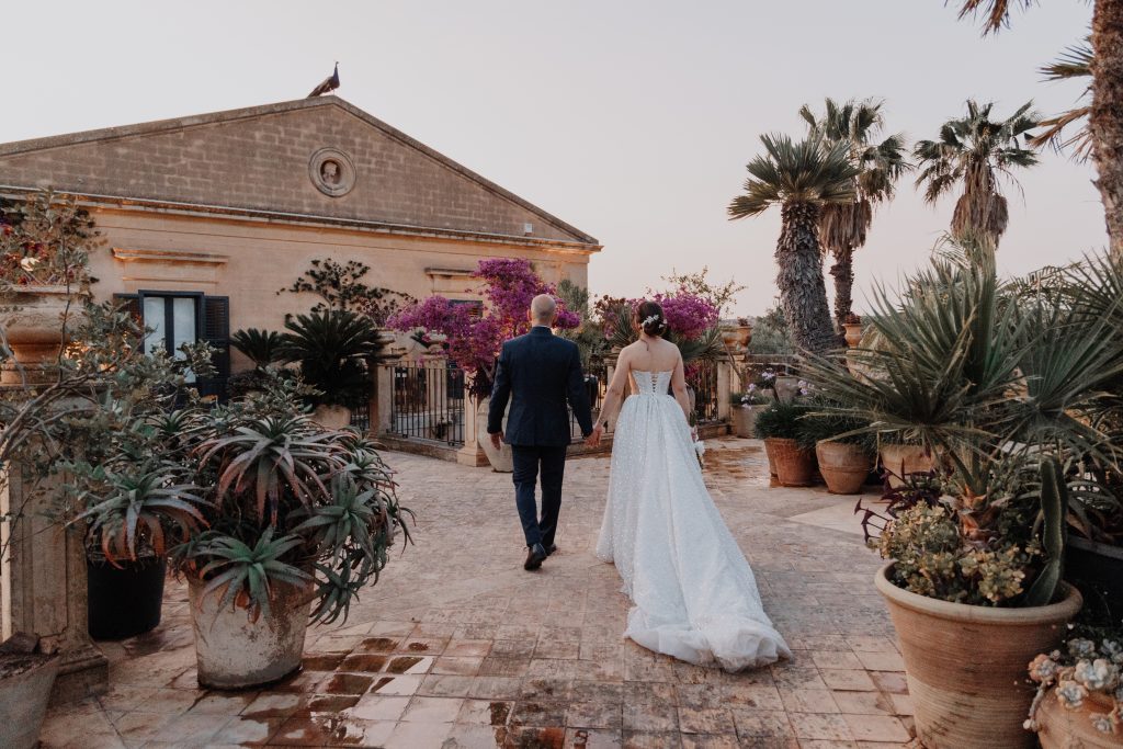 In questa foto di Vincenzo Pioggia si vede la coppia di sposi di spalle che si tiene per mano mentre cammina in un viale di una villa rustica. Intorno a loro piante in vaso, fiori rosa e palme tropicali 