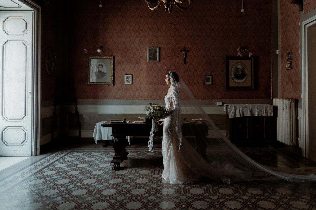 In questa foto di Vincenzo Pioggia si vede la sposa ritratta a figura intera di profilo. Si trova in un stanza di una casa antica, con pavimenti che richiamano motivi geometrici. 