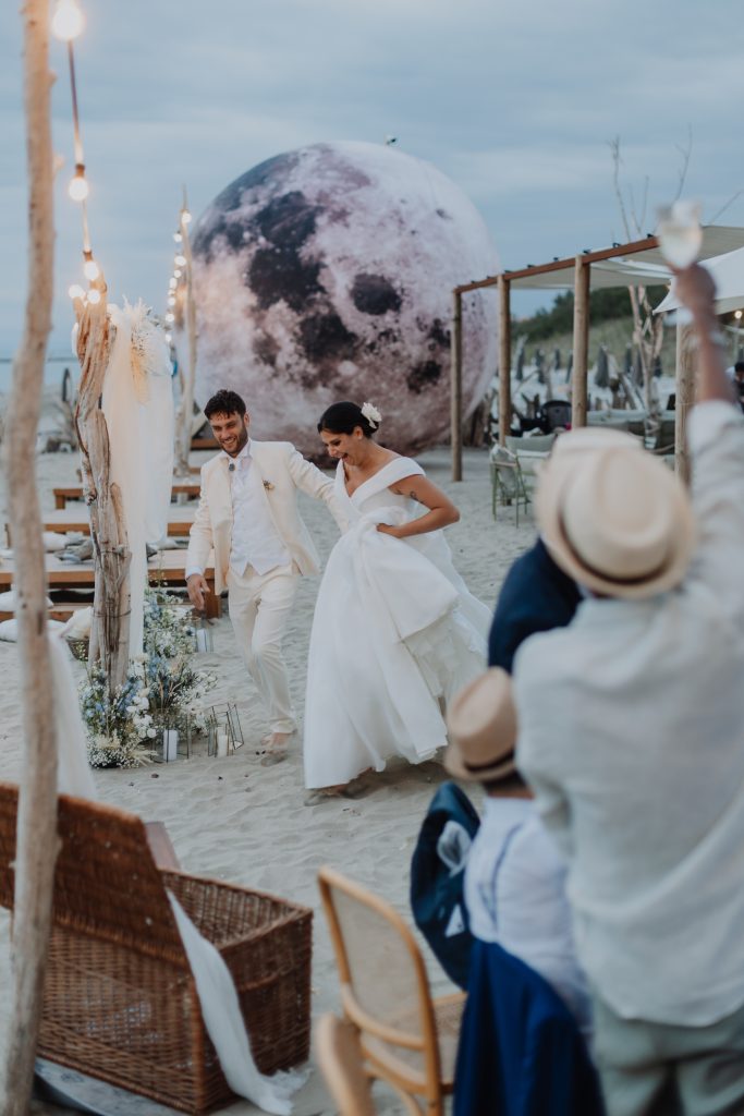 In questa foto di Vincenzo Pioggia si vede una coppia di sposi che cammina sorridente sulla sabbia durante un matrimonio in spiaggia. Lo sposo indossa un completo elegante di colore panna, mentre la sposa porta un abito da sposa ampio di colore bianco con scollo a V e fiore tra i capelli. Sullo sfondo si nota una scenografica luna gigante e luminosa, insieme a decorazioni naturali con fiori e rami. Alcuni invitati applaudono la coppia con calici alzati. L’atmosfera è gioiosa, rilassata e suggestiva.