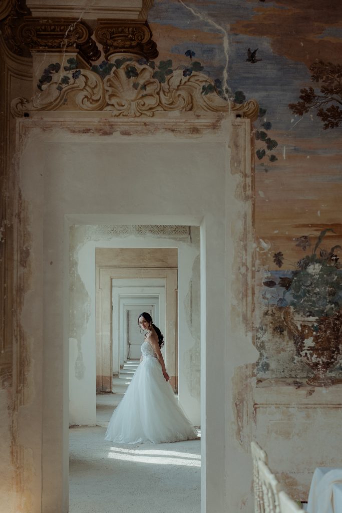 In questa foto di Vincenzo Pioggia si vede una sposa in piedi al centro di un lungo corridoio decorato, incorniciata da una serie di porte allineate che creano una prospettiva profonda e suggestiva. Indossa un abito bianco con corpetto ricamato e gonna ampia, e guarda indietro con grazia, voltandosi verso l'obiettivo. La luce naturale illumina la scena, valorizzando la delicatezza dell’abito e i dettagli degli affreschi sulle pareti. L’atmosfera è elegante, sospesa e senza tempo.