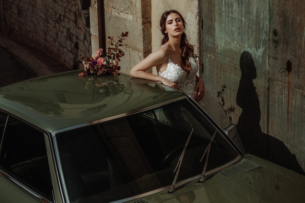 In questa foto di Vincenzo Pioggia si vede una sposa che esce da una macchina d'epoca, sul tettuccio della macchina è posato un bouquet di fiori di colore rosa. La sposa ha i capelli castani lunghi raccolti in una mezza coda
