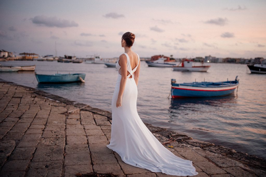 In questa foto di Vincenzo Pioggia si vede una sposa di spalle mentre cammina su un porticciolo. L'abito bianco ha uno scollo a incrocio sul retro e i capelli sono acconciati in uno chignon. 
Sullo sfondo il mare al tramonto e delle piccole barche 
