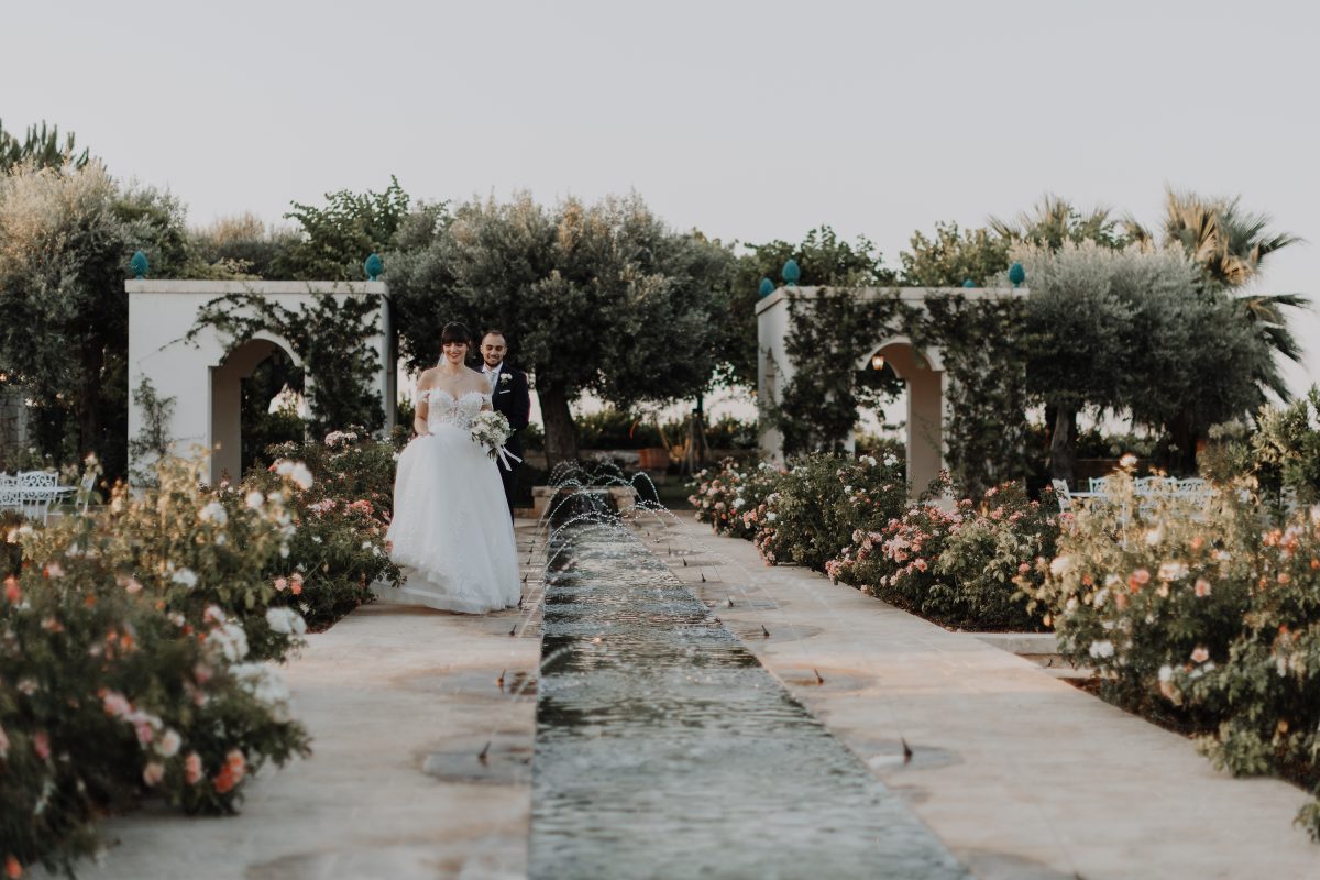 In questa foto di Vincenzo Pioggia si vede la coppia di sposi che cammina sorridendo verso il fotografo. SI trovano nel giardino di una villa e camminano su un viale pieno di piante e fiori con unna fontana 