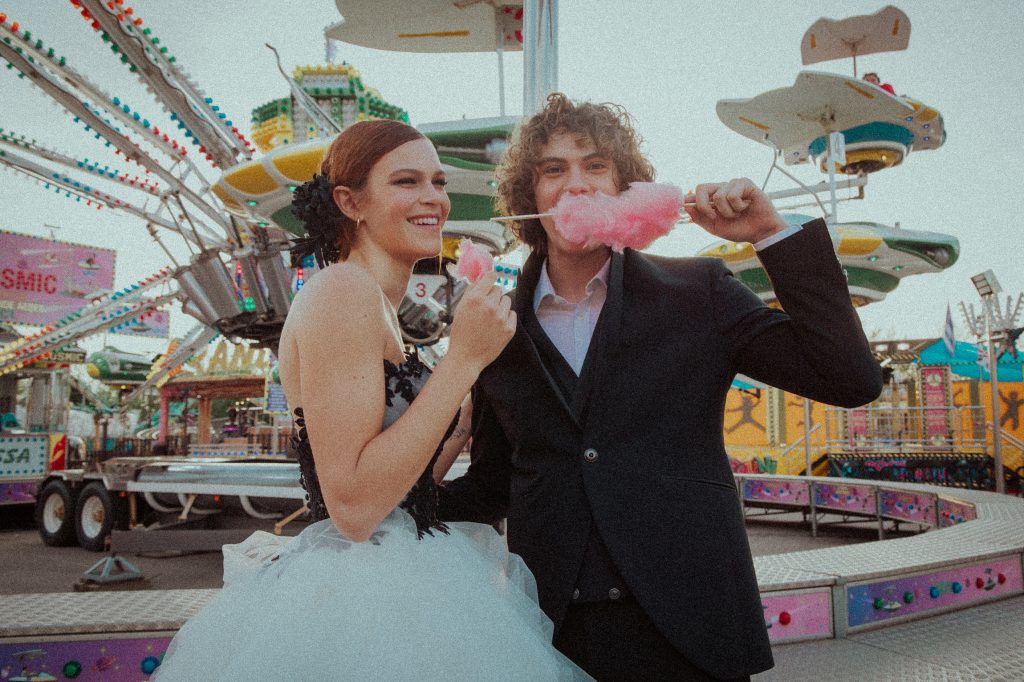 In questa foto di Vincenzo Pioggia si vede una coppia di sposi durante un servizio fotografico ambientato in un luna park coloratissimo mentre mangiano zucchero filato di colore rosa. Dietro di loro una giostra ottovolante.  