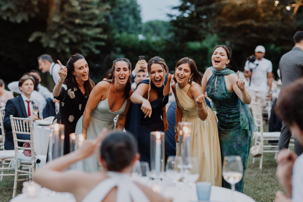 In questa foto di Vincenzo Pioggia si vede la sposa di spalle che sta per scattare una foto a cinque amiche durante il ricevimento di nozze. Le amiche in primo piano sorridono alla sposa. Da sinistra, la prima ragazza indossa un abito scuro a mezza manica, la seconda un abito scollato di colore azzurro carta da zucchero, la terza da sinistra ha un abito blu scuro. La quarta da sinistra ha un abito giallo chiaro con incrocio sul davanti, chiude la fila una ragazza mora con i capelli raccolti e un abito verde acqua simil maculato