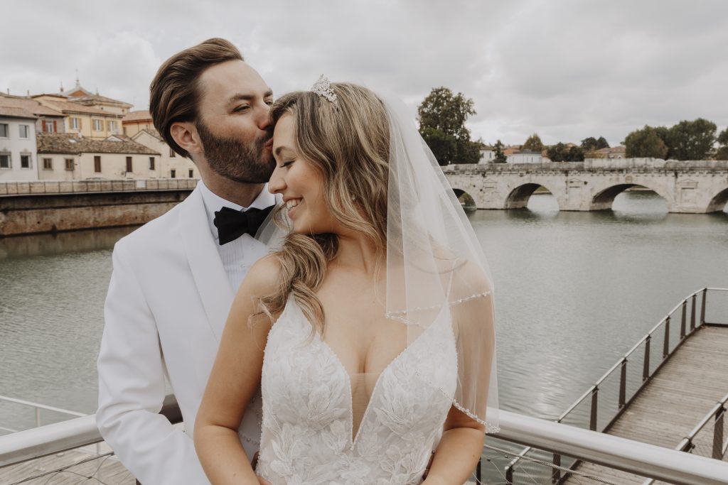 In questa foto di Vincenzo Pioggia si vede una coppia di sposi abbracciata su una passerella affacciata su un fiume. Lo sposo, in smoking bianco con papillon nero, bacia teneramente la fronte della sposa, che sorride con gli occhi chiusi. Lei indossa un abito bianco ricamato e un velo con perline, e tiene le mani intrecciate davanti a sé. Sullo sfondo si vede un antico ponte di pietra a cinque arcate e case colorate, sotto un cielo nuvoloso. L’atmosfera è romantica e serena.
