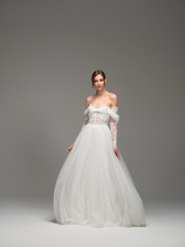In questa foto, una modella indossa uno degli abiti da sposa Maria Pia Creazioni 2026 modello ad A, in tulle, con scollo omerale e manicotti in pizzo floreale