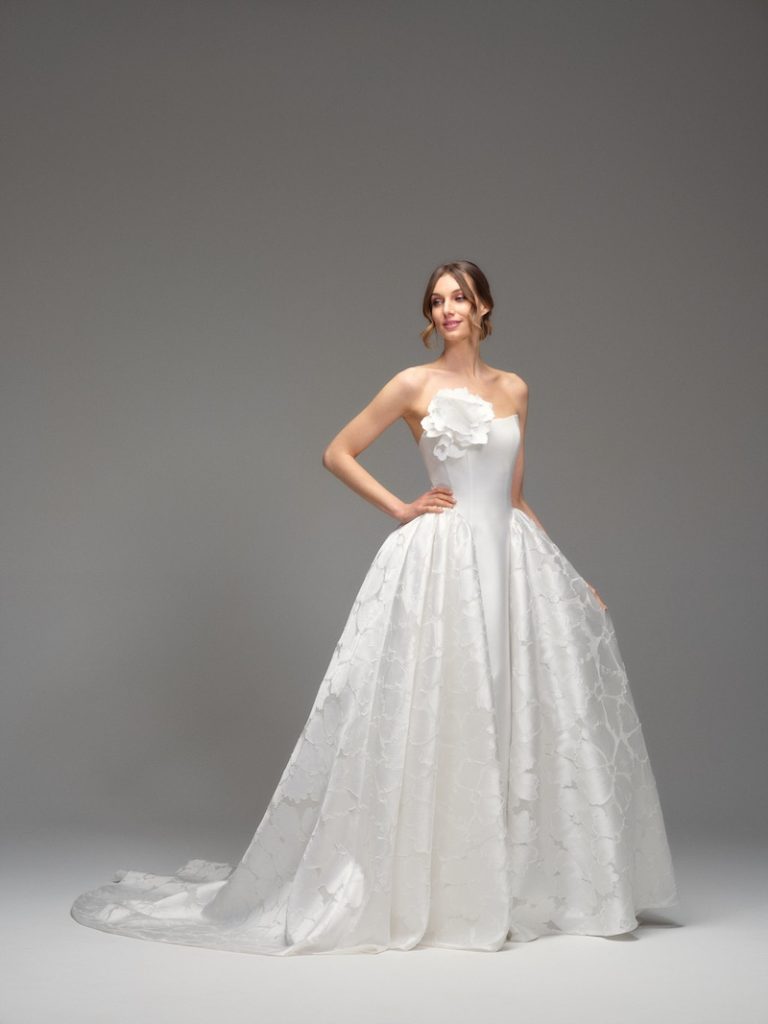 In questa foto, una modella indossa uno degli abiti da sposa Maria Pia Creazioni 2026 modello ampio, in mikado di seta, con sovragonna in tessuto marmorizzato e un maxi fiore 3D sul corpetto