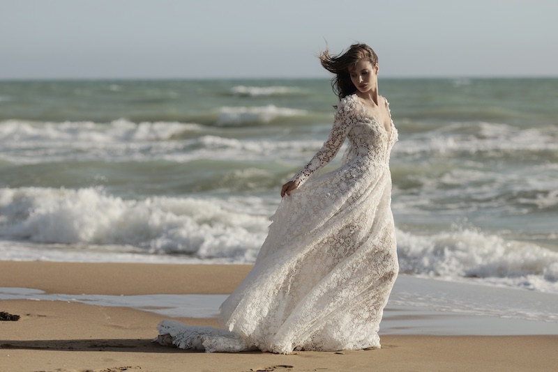 In questa foto, una modella posa camminando su una spiaggia davanti al mare mosso dal vento. Indossa un abito Luisa Sposa 2026 con pizzo floreale, maniche lunghe e scollo a V