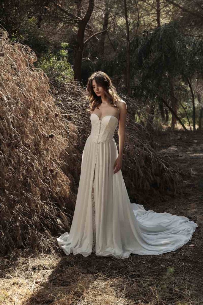 In questa foto, una modella posa in un giardino indossando un abito Luisa Sposa 2026 modello scivolato con spacco frontale, con. bustier e scollo a cuore
