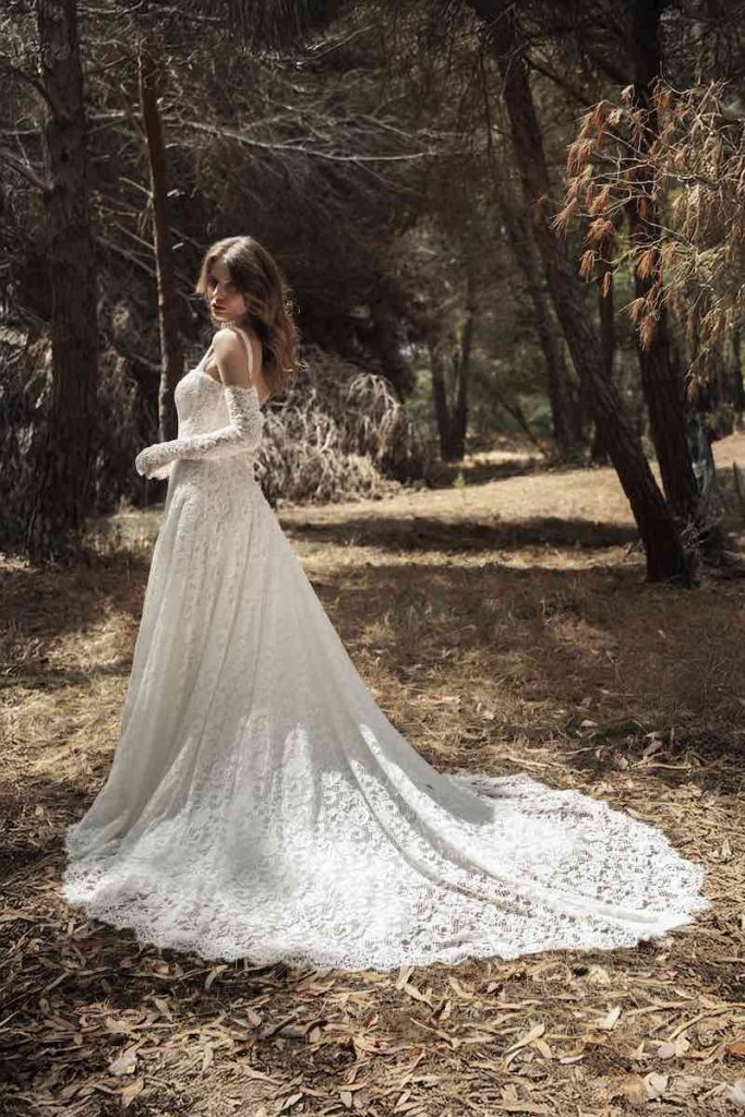 In questa foto, una modella posa di spalle in un giardino indossando un abito Luisa Sposa 2026 scivolato con lungo strascico interamente in pizzo floreale, con manicotti abbinati