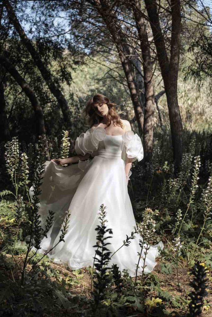 In questa foto, una modella posa in un giardino indossando un abito Luisa Sposa 2026, modello ad A in organza di seta, con manicotti con rouches e cintura drappeggiata