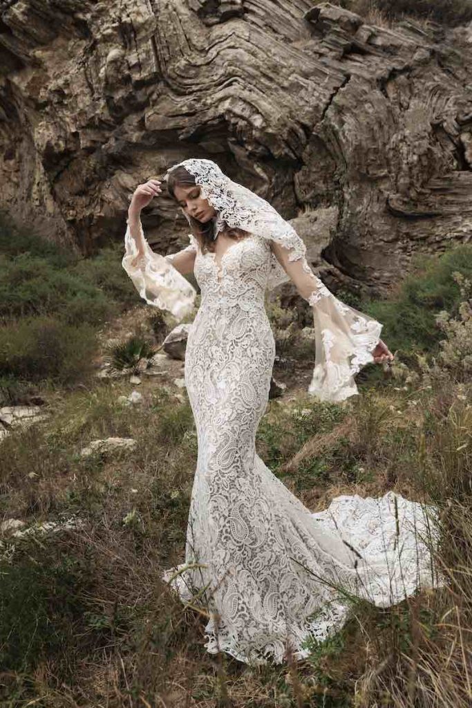 In questa foto, una modella posa su una roccia indossando un abito Luisa Sposa 2026 modello a sirena, con gonna in pizzo, maniche a campana e velo corto in pizzo