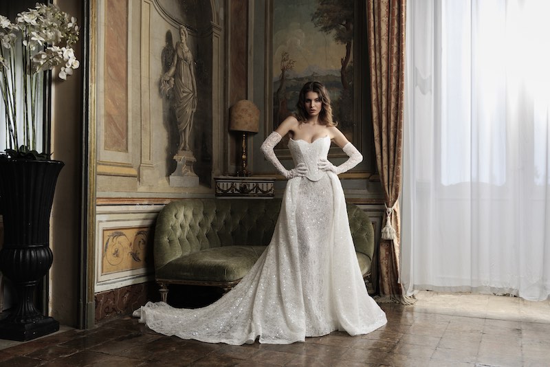 In questa foto, una modella posa nel salone di un palazzo nobiliare indossando un abito da sposa modello sirena con sopragonna e bustier staccabile e manicotti, Il tessuto è impreziosito da ricami d'argento