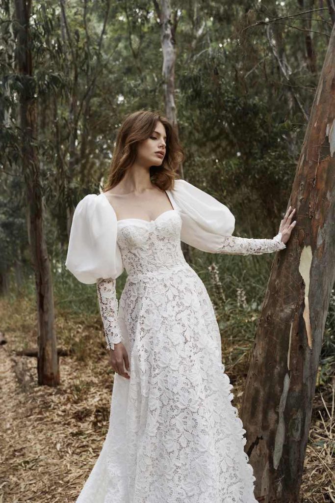 In questa foto, una modella posa in un giardino indossando un abito Luisa Sposa 2026, modello scivolato  interamente in pizzo macramè con maniche lunghe balloon