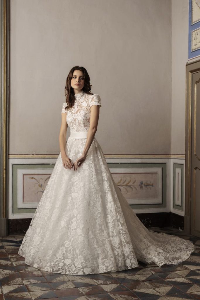 In questa foto, una modella posa nel salone di un palazzo nobiliare indossando un abito da sposa ad A Luisa Sposa 2026 in pizzo floreale con collo alto, maniche corte e cintura in vita