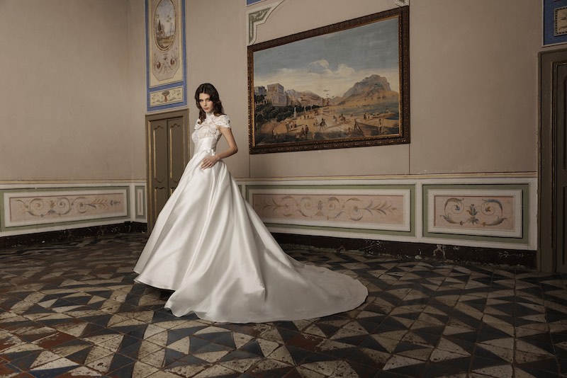 In questa foto, una modella posa nel salone di un palazzo nobiliare indossando un abito da sposa modello principessa con gonna in mikado di seta, bustier, maniche corte in pizzo e collo alto