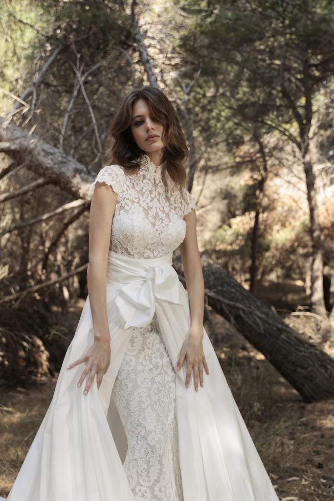 In questa foto, una modella posa in un giardino indossando un abito Luisa Sposa 2026, modello a sirena in pizzo floreale con collo alto e maniche corte e sopraggona in mikado fermato da un maxifiocco