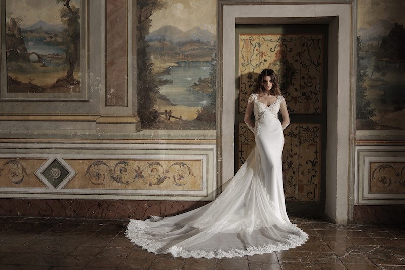 In questa foto, una modella posa nel salone di un palazzo nobiliare indossando un abito Luisa Sposa modello a sirena con maniche lunghe effetto illusion, scollo a cuore, lungo strascico in tulle con bordo ricamato