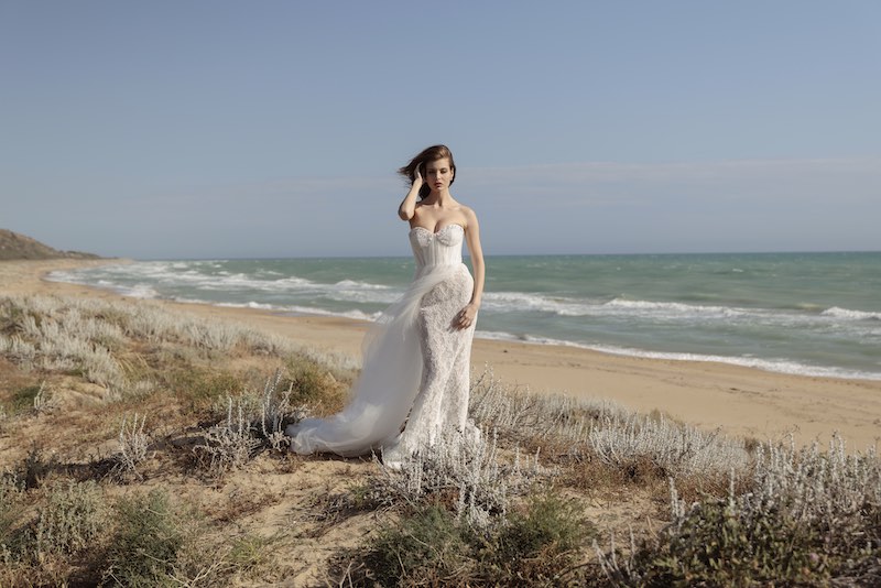 In questa foto, una modella posa su una spiaggia indossando un abito Luisa Sposa 2026 modello a sirena, con gonna in pizzo, sopragonna in tulle, bustier con scollo a cuore