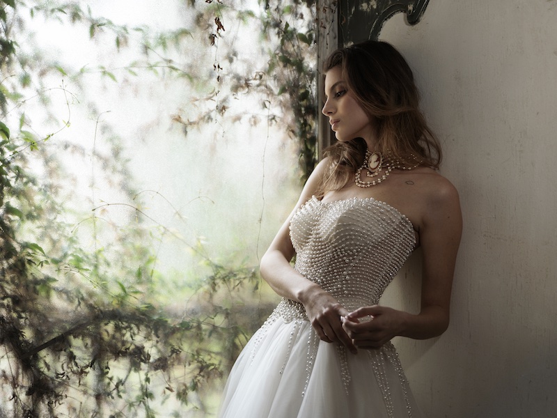 In questa foto, una modella posa guardando un giardino fiorito e indossando un abito da sposa con scollo a cuore Luisa Sposa 2026