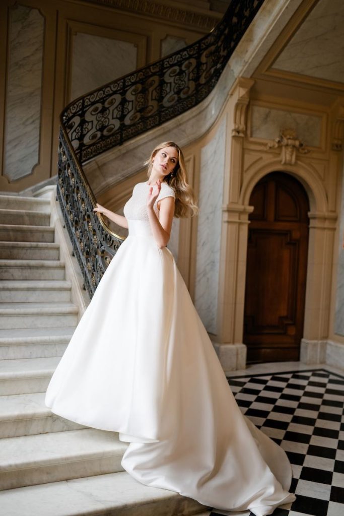 n questa foto, una modella posa sulla scalinata in marmo di una dimora storica indossando un abito da sposa dalla silhouette ad A, con maniche corte