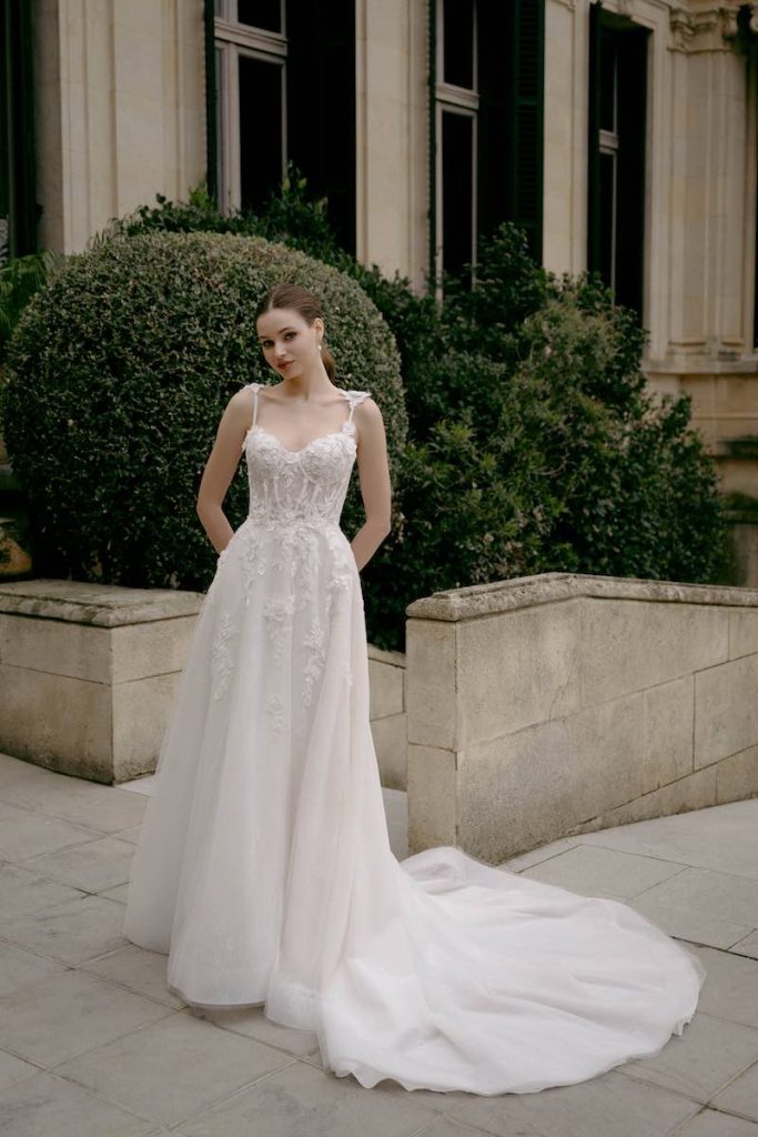 In questa foto, una modella posa nel giardino di una dimora storica indossando un abito da sposa Abella 2026, modello scivolato icon ricami di pizzo floreale che dal corpetto scendono ramage sul punto vita