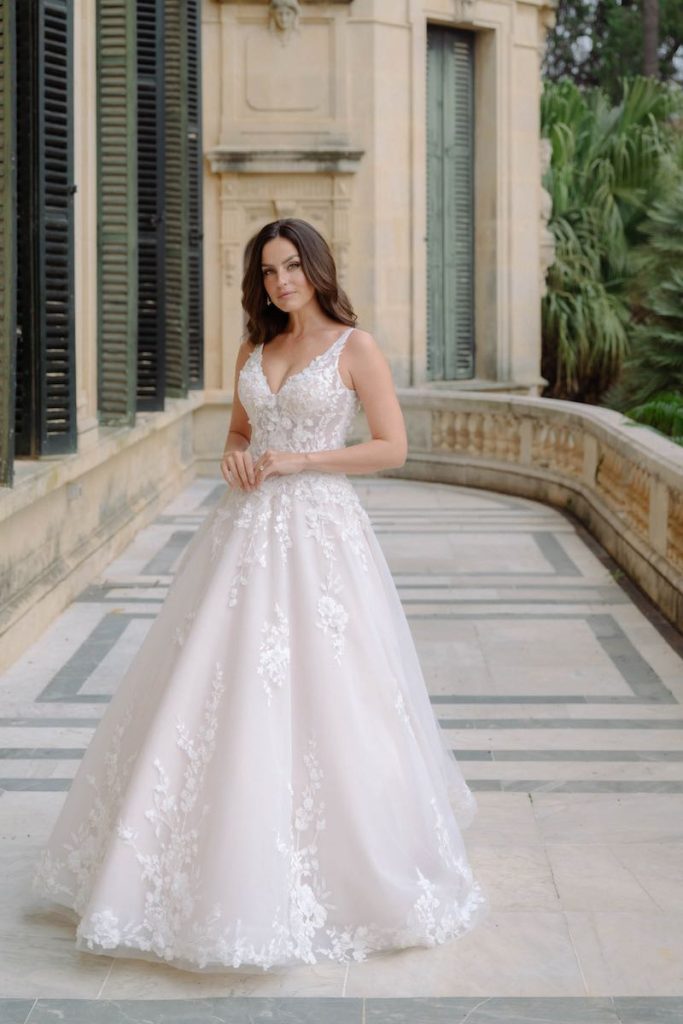In questa foto, una modella posa sulla balconata di una dimora storica indossando un abito da sposa ad A in tulle e inserti di pizzo floreale della collezione sposa Abella 2026