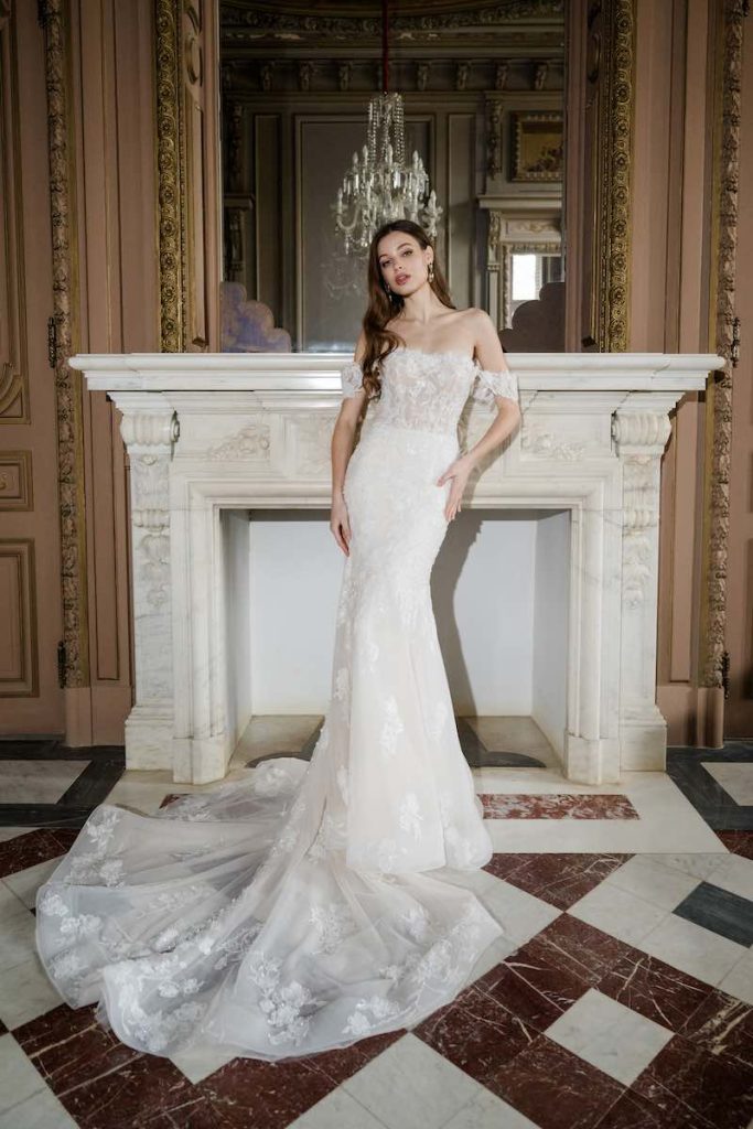 In questa foto, una modella posa nel salone di una dimora storica indossando un abito da sposa a sirena della collezione Abella 2026 con scollo off shoulder, ricami in pizzo floreale e bustier