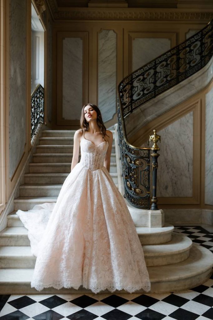 In questa foto, una modella posa sulla scalinata in marmo di una dimora storica indossando un abito da sposa Abella 2026 dalla silhouette ad A, con bustier a vista, scollo a cuore e in pizzo in nuance di rosa
