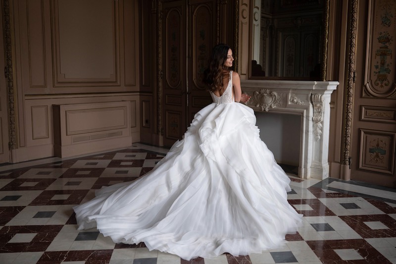 In questa foto, una modella posa di spalle nel salone di una dimora storica indossando un abito della collezione sposa Abella 2026 con lungo strascico, maxi balze e scollo sulla schiena