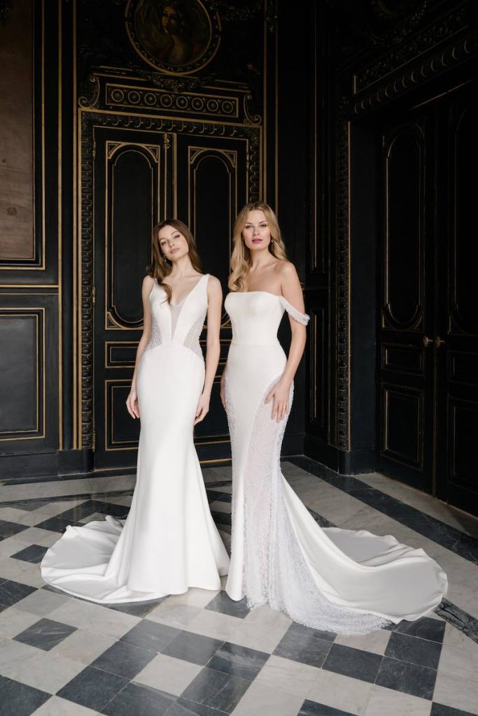 In questa foto, due modelle posano nel salone di una dimora storica indossando due abiti da sposa a sirena in cady di seta della collezione Abella 2026. Il modello a sinistra presenta dei dettagli cut out effetto illusion. Il modello a destra presenta uno scollo off shoulder e dettagli di perle tridimensionali sulle lunghezze